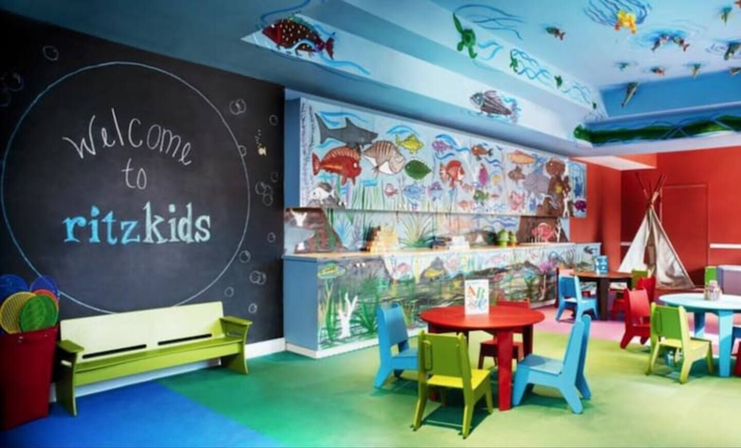 Zona infantil con un letrero de pizarra, sillas y mesas coloridas, y un mural de peces.