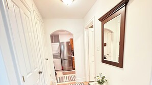 A hallway with white doors, a wooden floor, a bench, a mirror, and a kitchen in the background. - The Cozy Inn (Halifax)