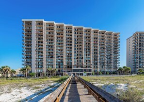 A high-rise hotel with multiple balconies, a wooden boardwalk leading to the entrance, and a clear blue sky. - Phoenix East 804 | Gulf Front | Sleeps 10 (Orange Beach)