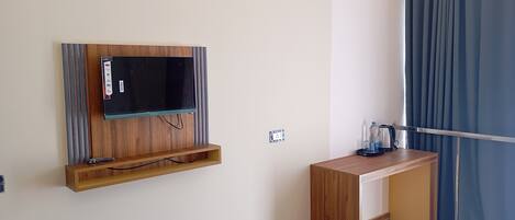 A room with a mounted TV, a wooden desk, a power outlet, and a window with curtains.