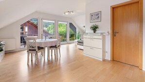 Une pièce spacieuse comprenant une table à manger, des chaises, un canapé, une porte en bois et de grandes fenêtres donnant sur un paysage verdoyant.
