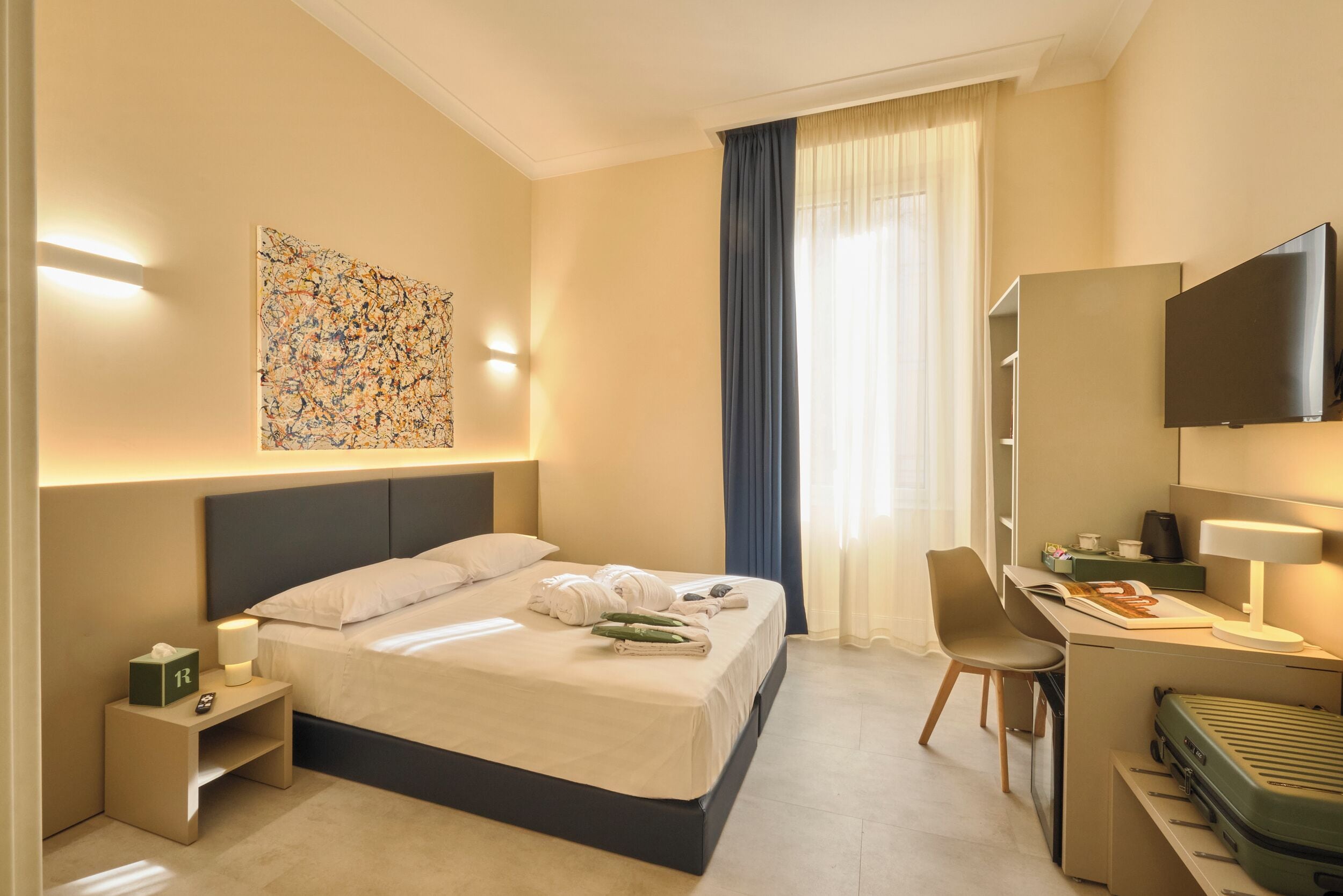 A hotel room with a bed, desk, chair, TV, and artwork on the wall.