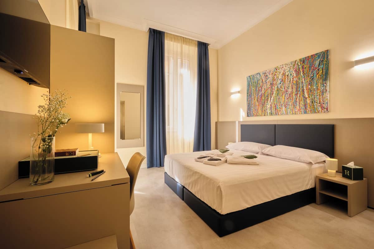 A hotel room with a bed, bedside table, desk, chair, and a painting on the wall.
