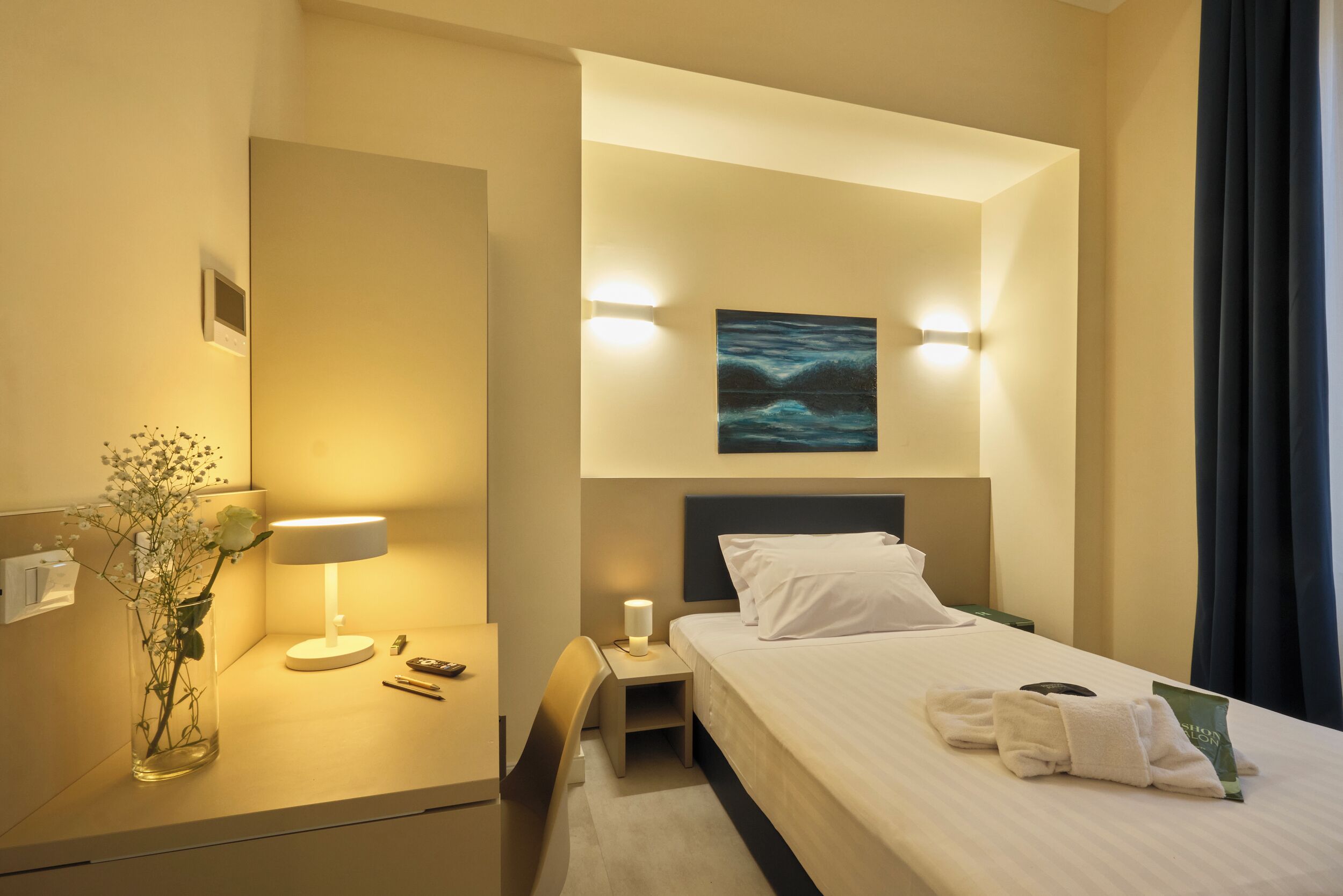 Una habitación de hotel con cama, mesita de noche, lámpara, jarrón con flores y un cuadro en la pared.