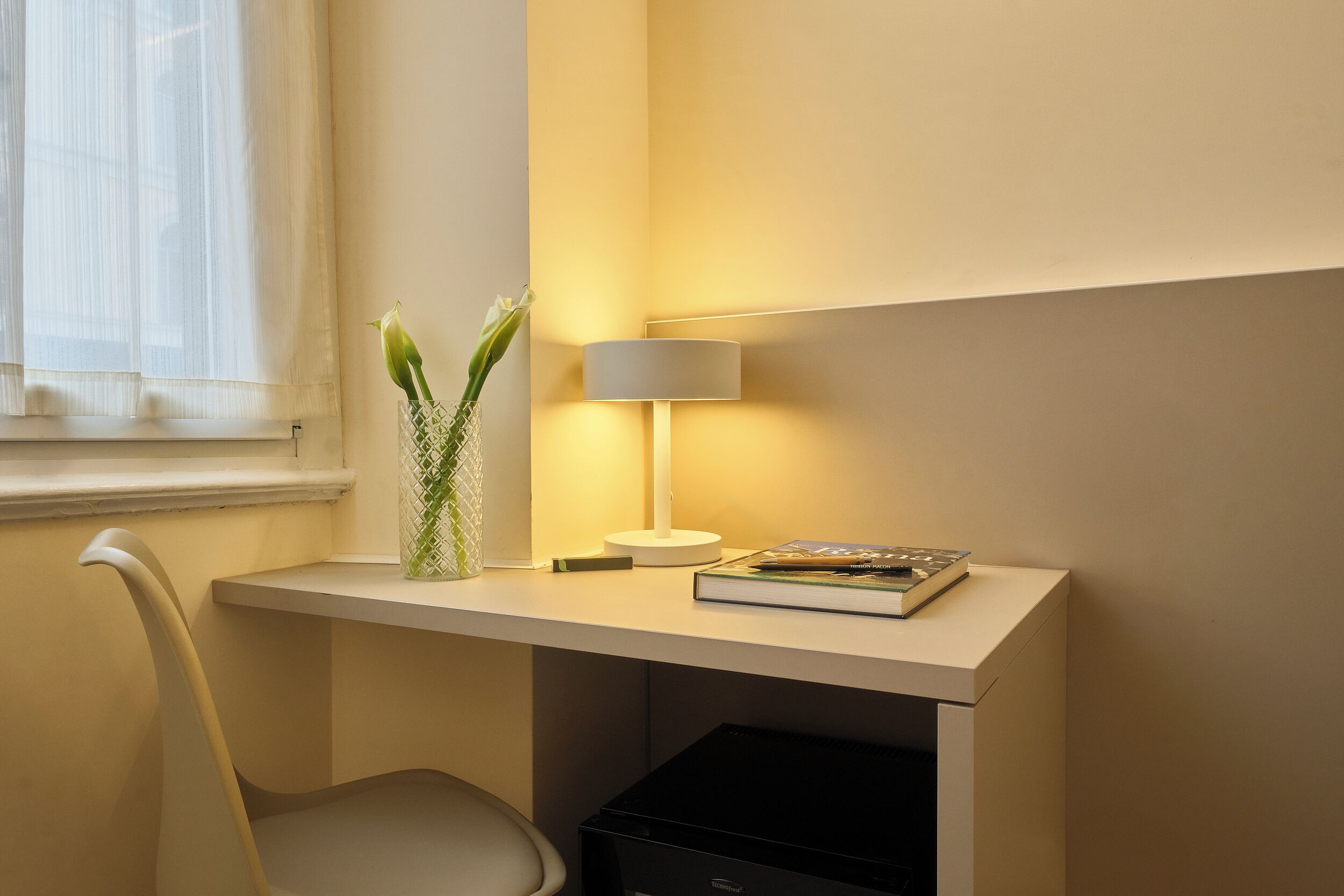 A desk with a lamp, a book, and a vase with flowers.