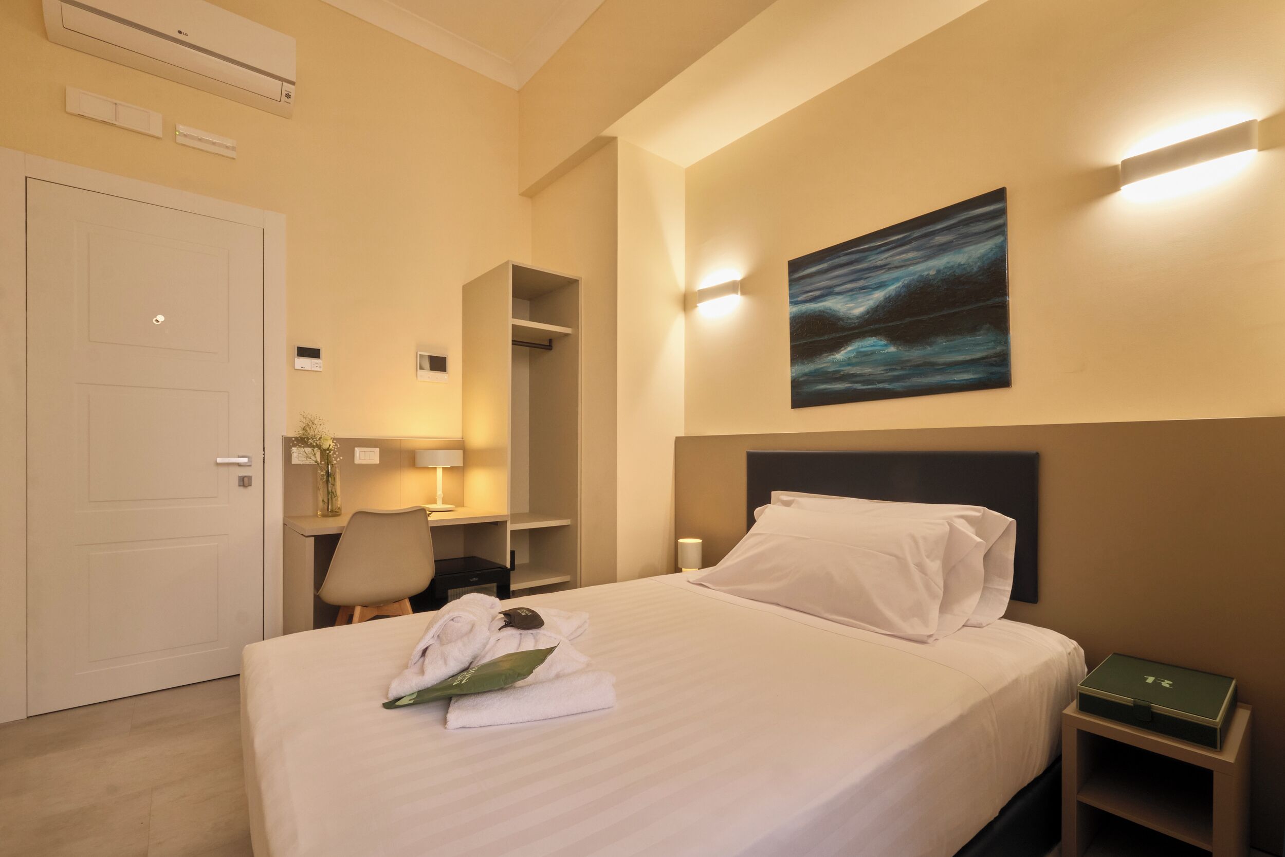 A hotel room with a bed, a desk, a chair, a nightstand, and a painting on the wall.