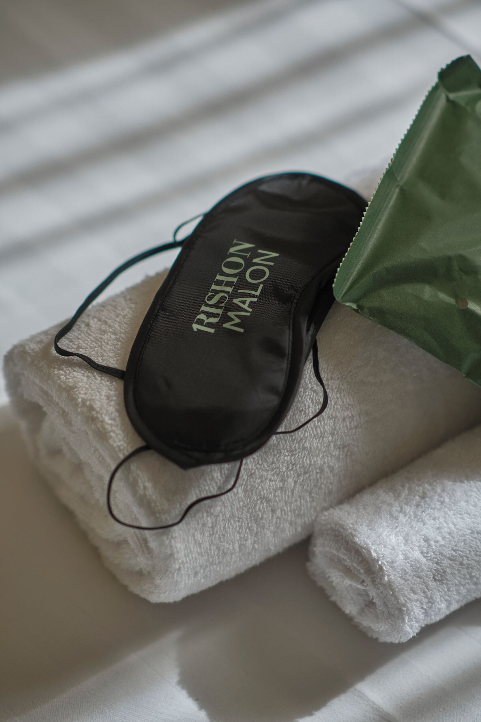 Een zwarte haarband met de tekst 'RISHON MALON', een witte handdoek en een groene plastic tas op een bed.