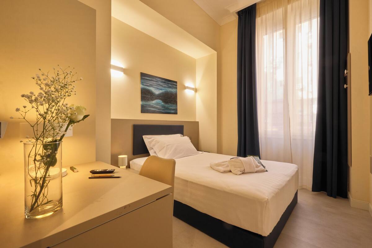 A hotel room with a bed, bedside table, lamp, and a painting on the wall.