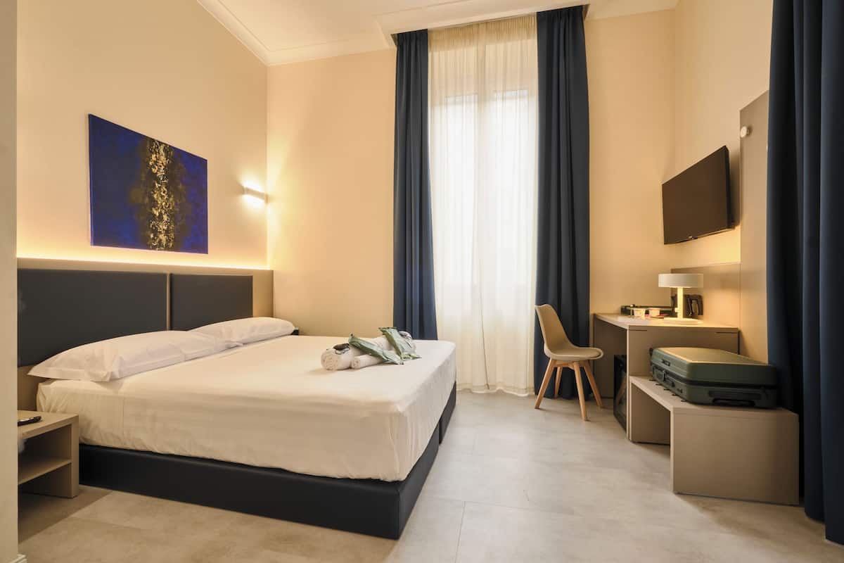 A hotel room with a bed, desk, chair, TV, and a painting on the wall.