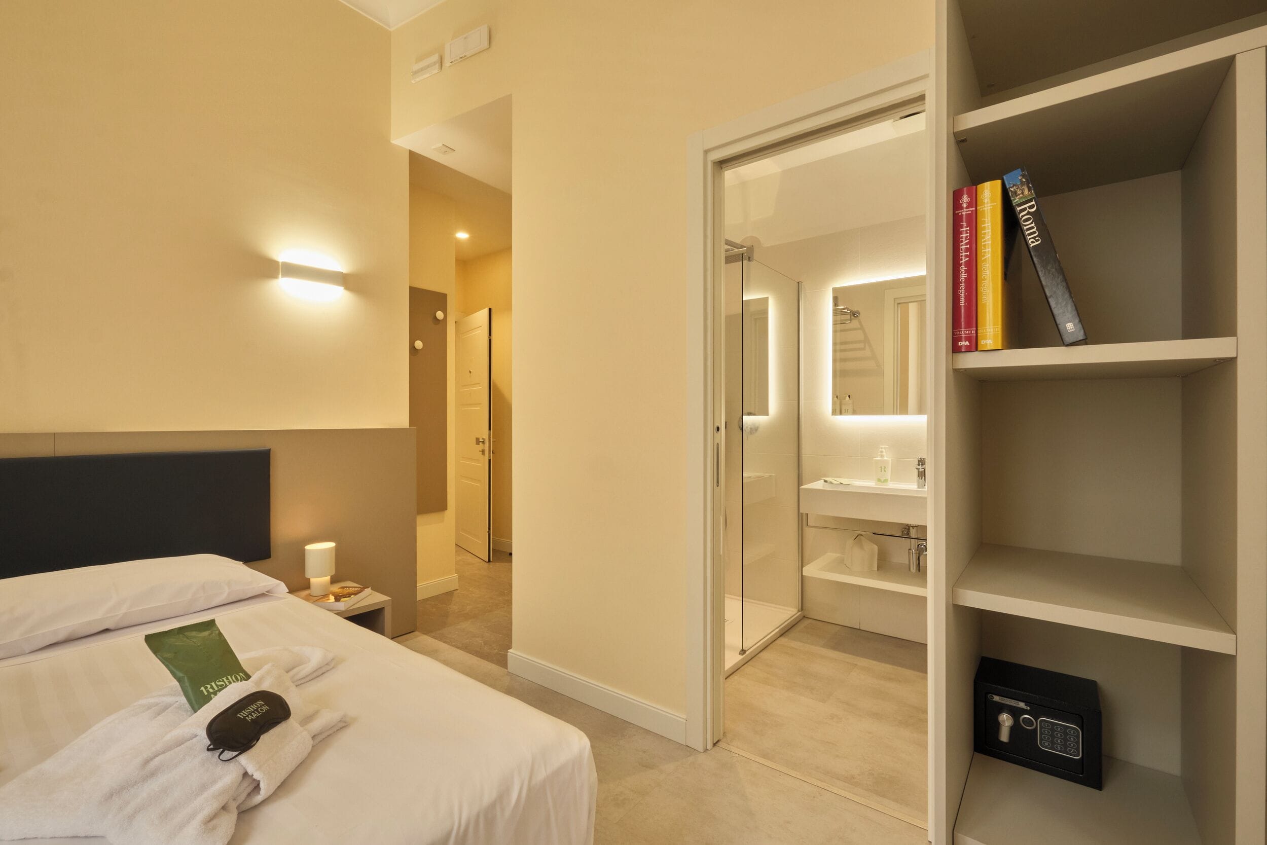 Una camera d'albergo con un letto, un comodino, un bagno con doccia in vetro e una libreria.