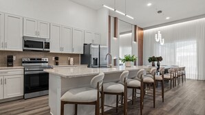 Una cocina moderna con armarios blancos, electrodomésticos de acero inoxidable, una isla central con taburetes altos y un área de comedor con un amplio ventanal.
