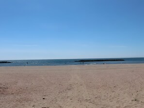 Une plage avec un ciel dégagé, une mer calme et des formations rocheuses visibles au loin.