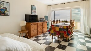 Una habitación con una cama, un televisor sobre un mueble de madera, una mesa de comedor con sillas y un ventanal con vistas.