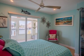 A bedroom with a bed, a chair, a ceiling fan, a door leading outside, and wall decorations including a fish and flowers. - Island Style Waterfront Retreat w Private Dock (Saint James City)