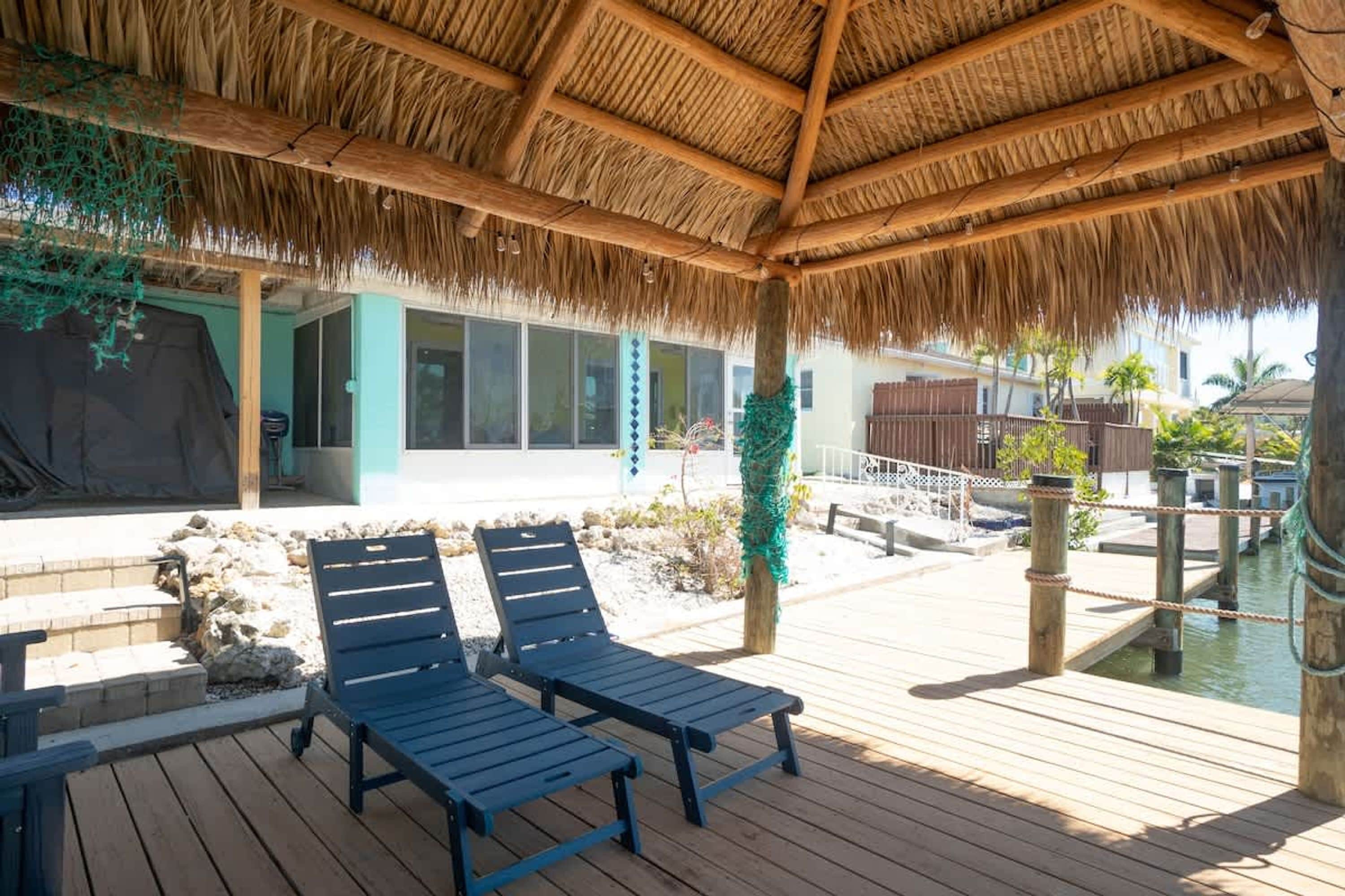 A wooden deck with two lounge chairs, a thatched roof, a pool, and a building with large windows.