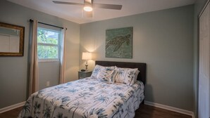 Une chambre à coucher avec un lit, un ventilateur de plafond, une fenêtre avec des rideaux, une lampe fixée au mur et un tableau.