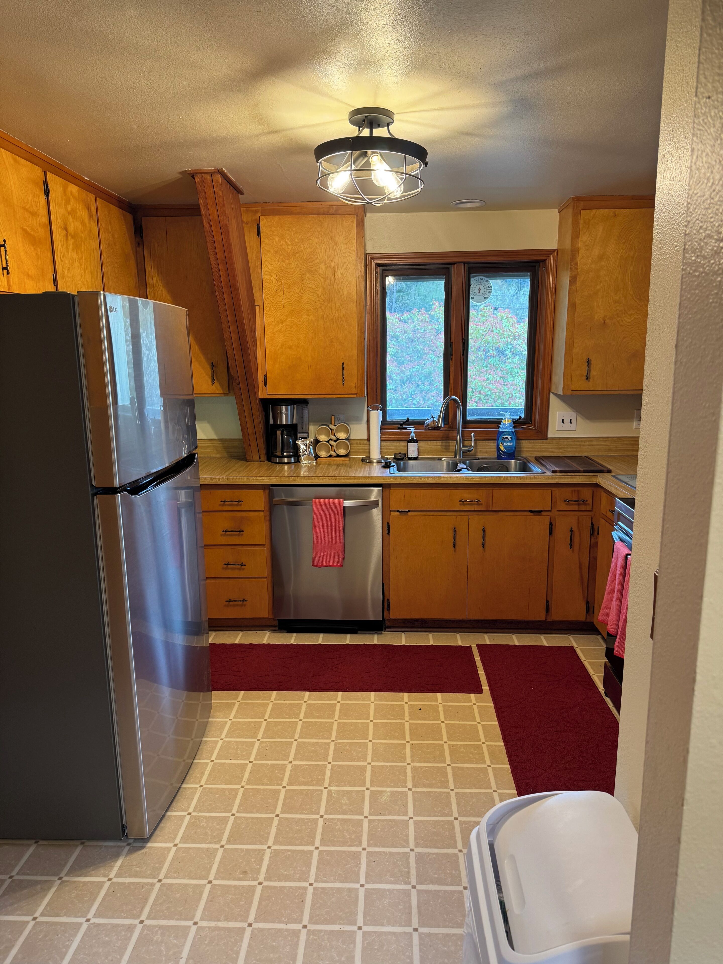 A kitchen with wooden cabinets, a stainless steel refrigerator, a window, and a red rug.