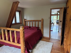 A bedroom with a wooden bed frame, a red bedspread, and a view through an open door to a dining area.