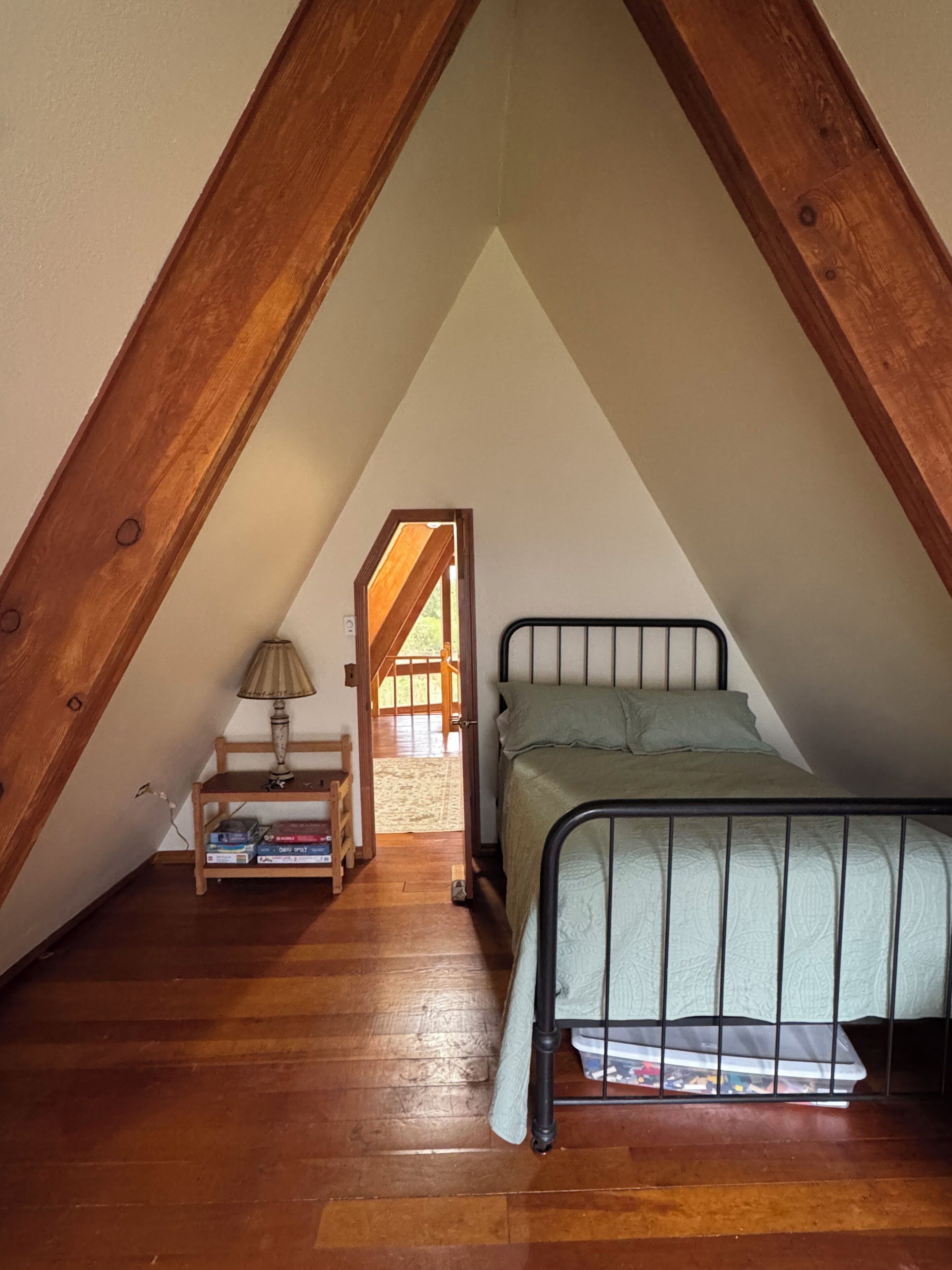 Un dormitorio en el ático con techo inclinado, cama, mesita de noche, lámpara y estantería.