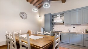 A dining area with a wooden table and white chairs, a kitchen with grey cabinets, and a wall clock.