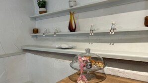 Ett kök med vita väggar, hyllor med dekorativa föremål, en bänkskiva i trä och ett glasskåp med bakverk.