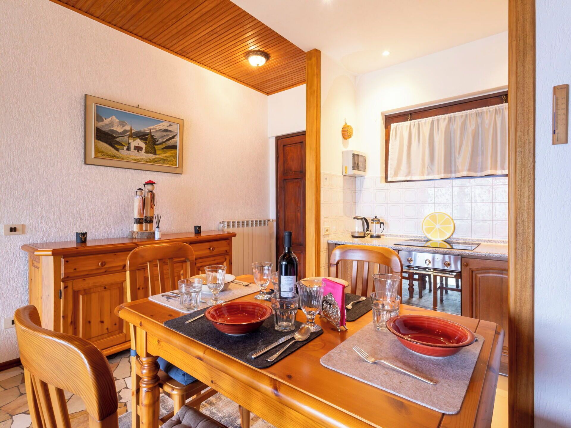 A dining area with a wooden table set for four, a kitchen in the background, and a painting on the wall.