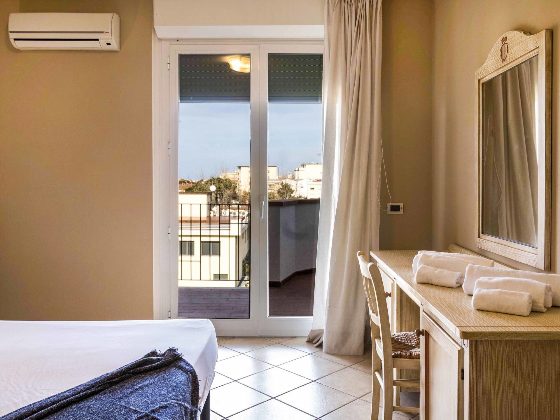 Une chambre d’hôtel avec un lit, un bureau doté d’un miroir, une salle de bain avec douche et une vue sur les bâtiments environnants.