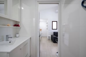 Ein modernes Badezimmer mit Waschbecken, Spiegel und Schränken in Weiß. Im Hintergrund ist ein kleiner Hocker zu sehen.