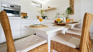 Eine moderne Küche mit weißen Schränken, einem Backofen, einem Holzschild mit der Aufschrift 'KITCHEN' und einem Essbereich mit Korbstühlen und einem weißen Tisch.