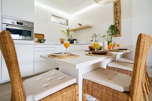 A modern kitchen with white cabinetry, an oven, a wooden sign reading 'KITCHEN', and a dining area with wicker chairs and a white table. - Royal Blue San Agustin (San Bartolomé de Tirajana)
