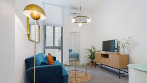 Un salon moderne avec un canapé bleu, une table basse ronde en verre, un téléviseur posé sur un meuble en bois et un luminaire suspendu.