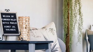 A room with a bed, a bedside table with a sign, a lamp, and hanging plants. - Charming Garden Route Villa, Nature View and BBQ Bliss (Mossel Bay)
