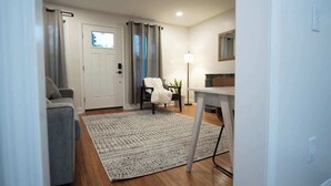 Una habitación con un sofá gris, una mesita, una silla, una lámpara y una puerta con cortinas.