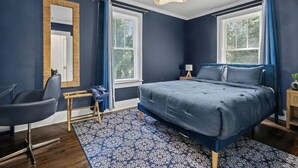 Un dormitorio con una cama, una silla, una mesita de noche, una lámpara y una ventana con cortinas.