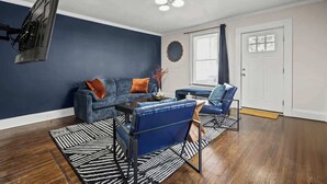 Una sala de estar con una pared de acento azul oscuro, piso de madera, un sofá azul con cojines anaranjados, una mesa de centro y un televisor montado en la pared.