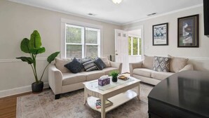 A living room with a beige sofa, a coffee table, a potted plant, and a door leading to another room. - Sleeps 10 - Plaza Midwood - Walk to Restaurants (Charlotte)