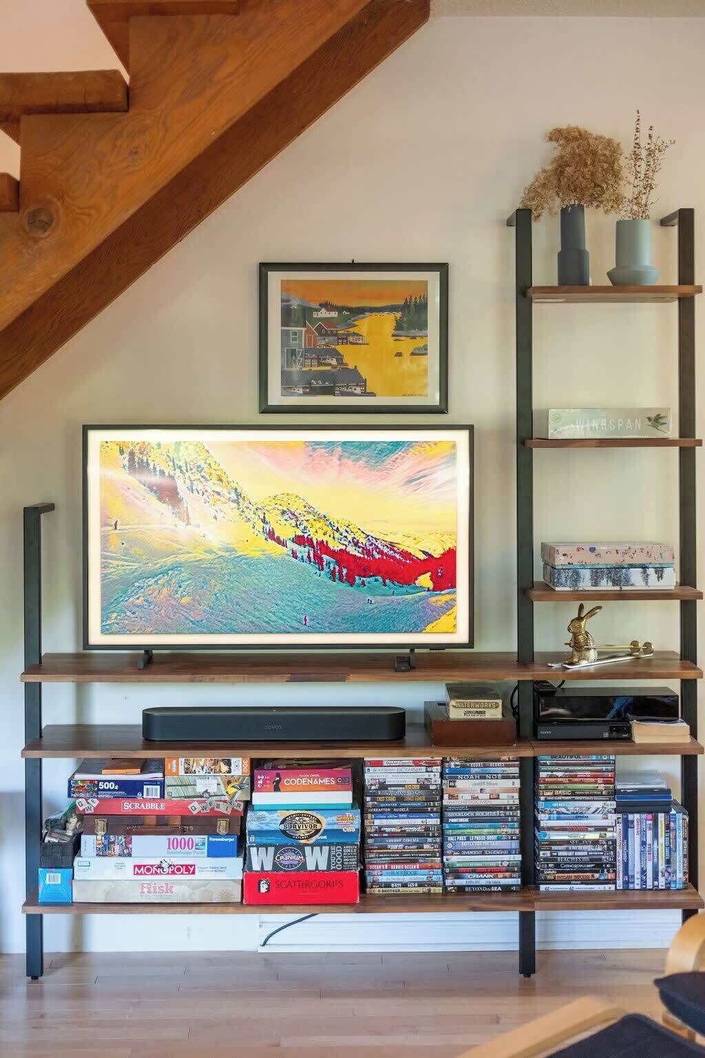 Una sala de estar con una televisión de pantalla plana, una estantería llena de juegos de mesa y un cuadro enmarcado sobre la televisión.