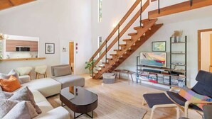 A spacious living area with a wooden staircase, a sofa set, a coffee table, and a bookshelf with books and decorative items.