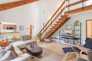 A spacious living area with a wooden staircase, a sofa set, a coffee table, and a bookshelf with books and decorative items.