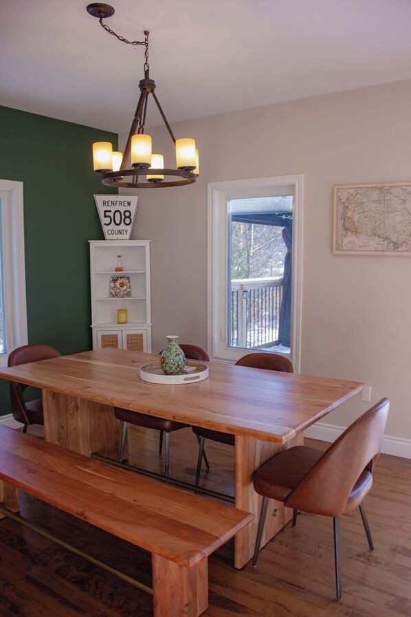 A dining area with a wooden table, benches, a chandelier, a window with a view, and a sign indicating the location.