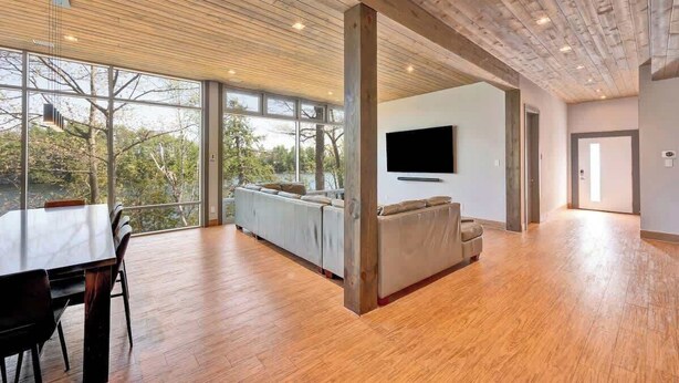 A spacious living area with a wooden ceiling, a large window offering a view of trees, a sofa, and a flat-screen TV mounted on the wall. - Lake House Wood Sauna & Hot Tub (Portland)