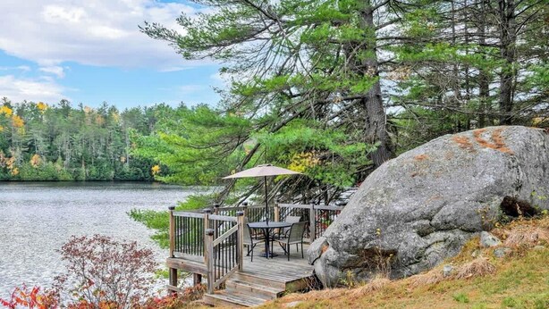 A lakeside deck with a table and chairs, a large rock, and a wooden railing. - KilDrummy Cottage with Sauna (Calabogie)