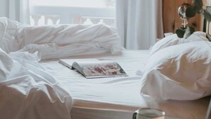 A neatly made bed with a magazine, a cup, and a candle on a table.