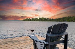 Une jetée en bois avec une chaise et un verre de vin, d’où l’on peut admirer un lac paisible et un ciel au coucher du soleil.