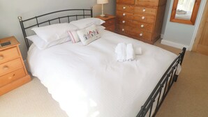 Een keurig opgemaakt bed met witte lakens, een houten dressoir met lades, een bedlampje en een spiegel aan de muur.