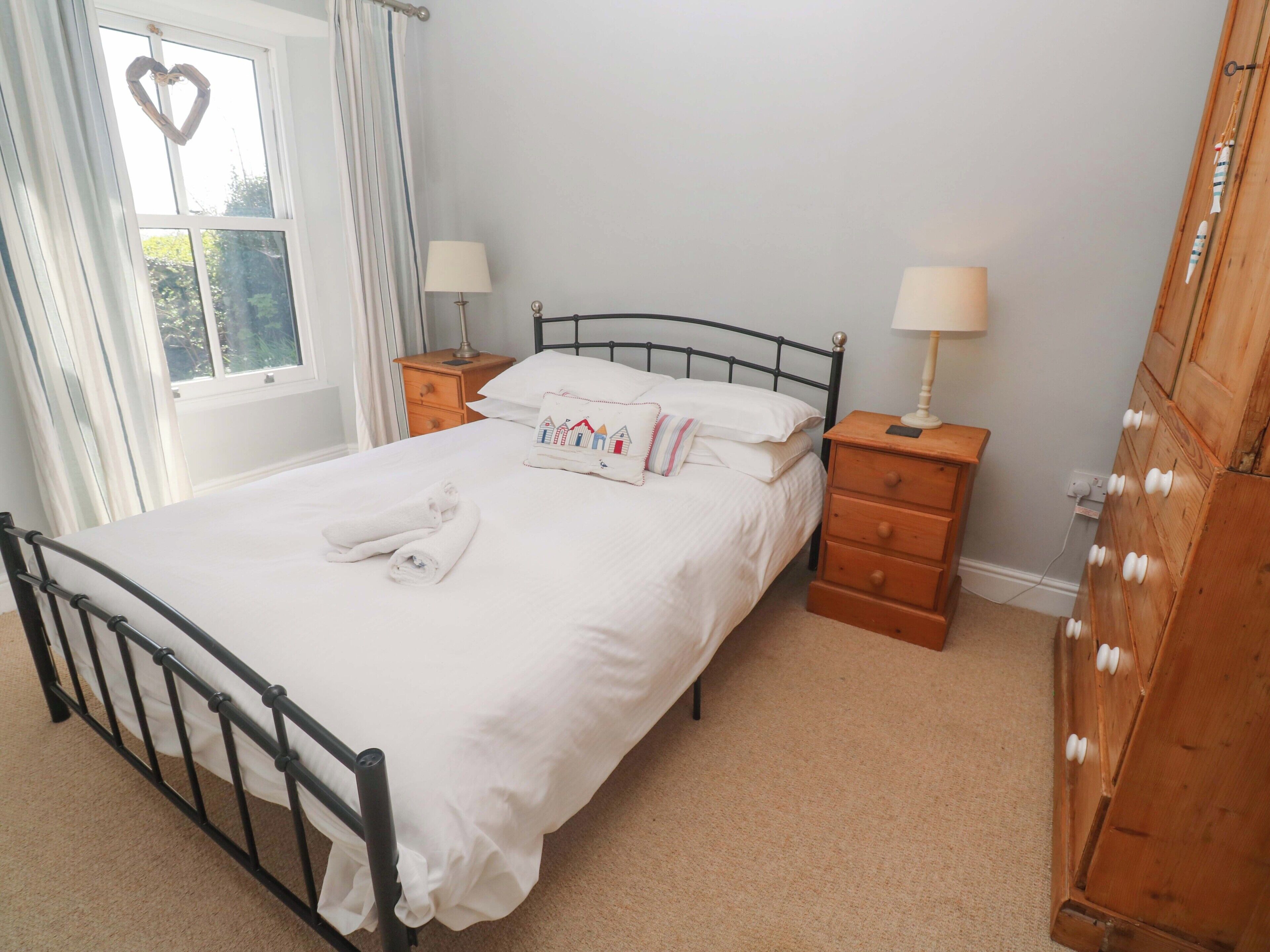 Uma cama bem arrumada com lençóis brancos, duas mesas de cabeceira com abajures, um aparador de madeira e uma janela com cortinas.