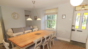 Een eetruimte met een houten tafel en witte stoelen, een klok aan de muur en een raam met gordijnen.