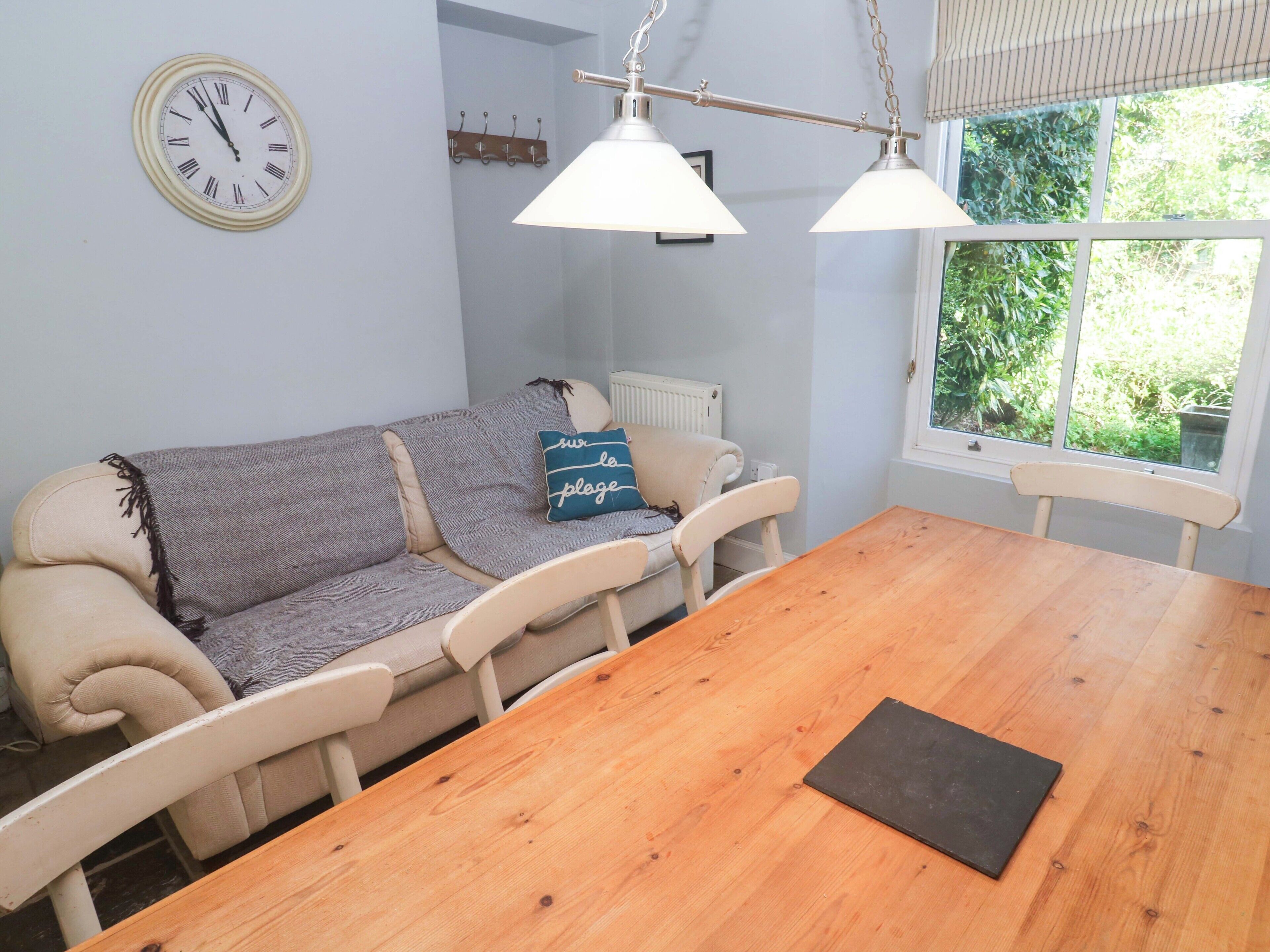 Ein Essbereich mit einem Holztisch, weißen Stühlen, einer Couch mit Kissen, einer Wanduhr und einem Fenster mit Grünpflanzen draußen.