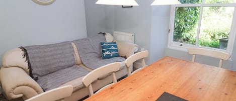 Ein Essbereich mit einem Holztisch, weißen Stühlen, einer Couch mit Kissen, einer Wanduhr und einem Fenster mit Grünpflanzen draußen.