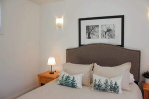 Un dormitorio con cama, almohadas, mesita de noche, lámpara y cuadros en la pared.